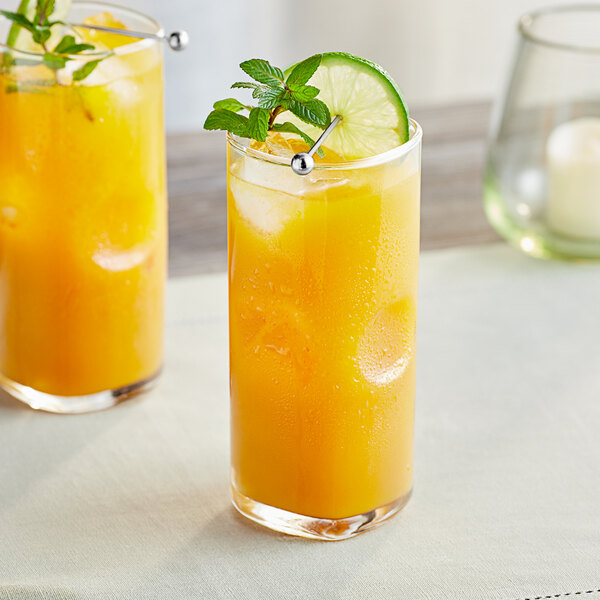 A glass of yellow liquid with a lime and mint garnish next to a glass of yellow liquid with a lime and mint leaf.